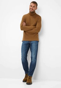 Maglietta a maniche lunghe, colore marrone, con colletto alto, abbinata a jeans blu e stivali marroni con lacci. La maglietta è aderente, con una texture liscia e una silhouette slim.