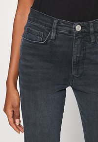Jeans in denim scuro con una vestibilità aderente, dotati di chiusura a bottone, passanti per cintura e tasche frontali. Il tessuto ha una texture liscia.