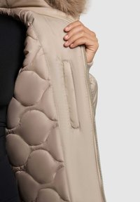 Beige quiltad jacka med slät yta, som har dragkedjestängning, krage med pälsdetalj och en sidoficka på ärmen.