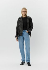 Chaqueta de motociclista de cuero negro con cremallera asimétrica, herrajes plateados y bolsillo de parche. Se lleva con una camiseta negra y jeans rectos de color azul claro.