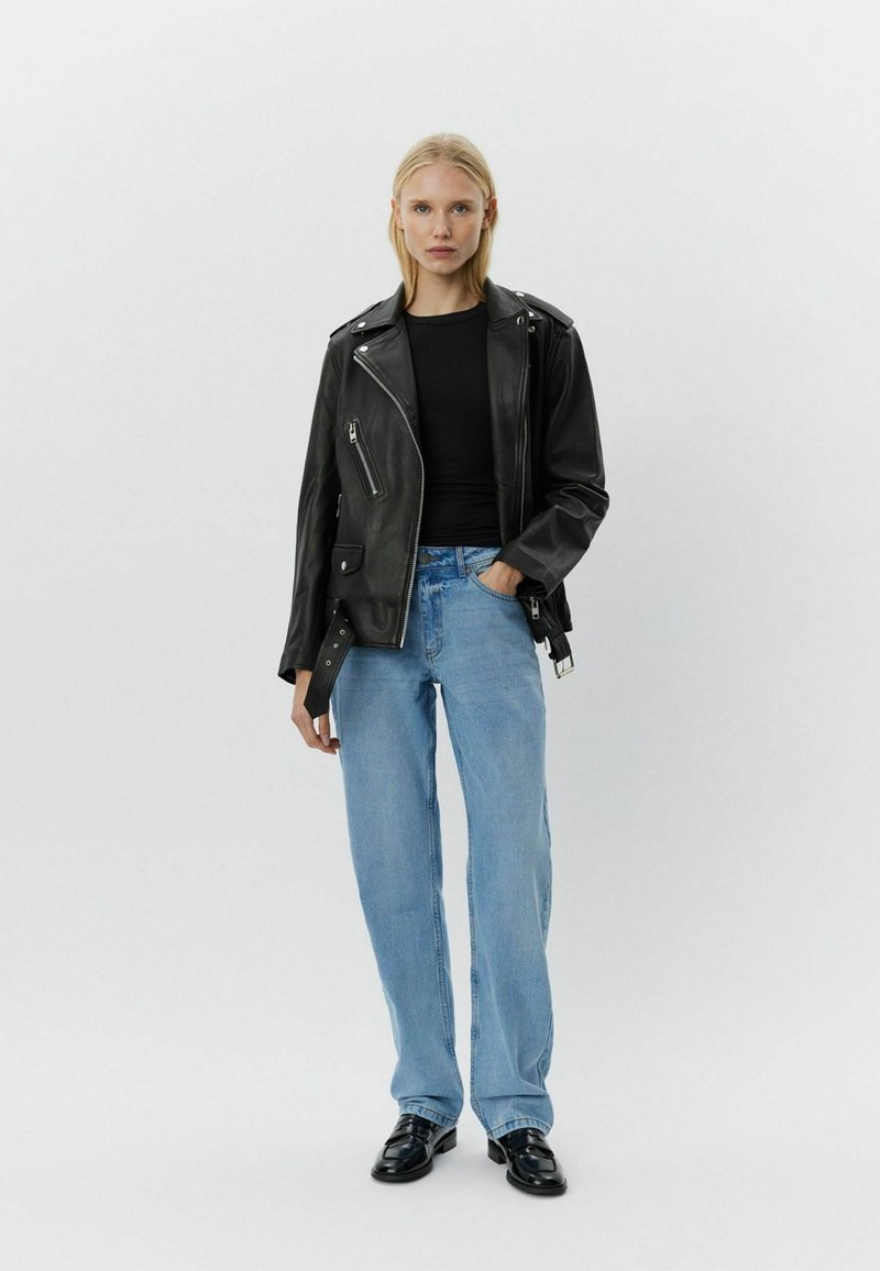 Chaqueta de motociclista de cuero negro con cremallera asimétrica, herrajes plateados y bolsillo de parche. Se lleva con una camiseta negra y jeans rectos de color azul claro.