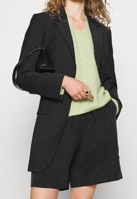 Femme portant un blazer noir et un short, un pull en maille vert clair, tenant un petit sac à main noir sous le bras, avec des bagues aux doigts.