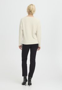 Maglione a coste crema con vestibilità ampia, abbinato a pantaloni neri slim-fit e stivali neri in vernice alla caviglia, visto da dietro.