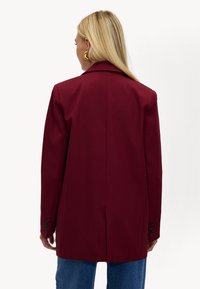 Bordeaux blazer met een gestructureerd ontwerp, enkel vent, en drie-knopen manchetten. Zachte stof met een getailleerde pasvorm, weergegeven van achteren.