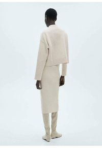 Hellbeige, taillierte Strickjacke und figurbetonte Midirock, beide aus weichem Strickmaterial, kombiniert mit knielangen beigen Stiefeln.