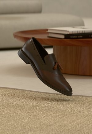 Mocasín de cuero marrón oscuro con borlas flotando sobre una alfombra beige tejida, con una mesa redonda de madera y libros apilados en el fondo.