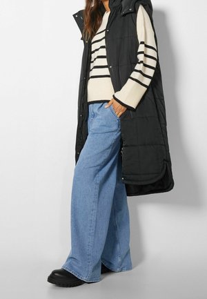 Vrouw met een zwart doorgestikt lang vest over een witte trui met zwarte strepen, wijde blauwe jeans en zwarte schoenen.