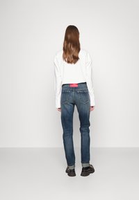 Jeans de mezclilla azul claro con corte recto, bolsillos traseros visibles y una etiqueta roja en la cintura. Combinados con una camisa blanca de manga larga.