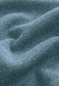 Gros plan sur un tissu bleu-gris doux et texturé avec un motif de tricot visible et de légers plis créant des ombres et des reflets subtils.
