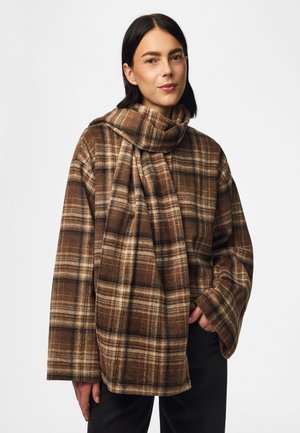 Femme aux cheveux noirs et raides porte un manteau oversize à carreaux marron et beige avec une écharpe assortie, posant avec une main dans la poche.