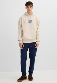 Joven de pie, vestido con una sudadera con capucha beige con texto azul, pantalones deportivos azul marino y zapatillas marrones sobre un fondo claro y liso.