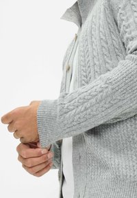 Graue Strickjacke mit einem Zopfmuster, gerippten Bündchen und Holzknöpfen. Nahaufnahme des Arms und der Stoffstruktur.