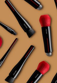 Schwarze Make-up-Pinsel mit roten Akzenten, die in unterschiedlichen Formen und Borstentexturen angeordnet sind, auf einem beige Hintergrund.