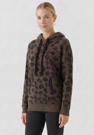Junge Frau mit blondem Haar trägt einen braunen Kapuzenpullover mit Leopardenmuster, schwarzen Kordeln und schwarze Hose, stehend vor einem einfarbigen Hintergrund.