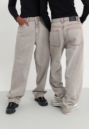 Dwie osoby ubrane w luźne jasnoszare dżinsy z czerwonym przeszyciem oraz czarne bluzki z długim rękawem, jedna stoi przodem z ręką w kieszeni, druga tyłem.