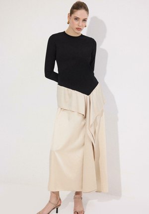 Γυναίκα με τα μαλλιά της σε κότσο, που φοράει χρυσά σκουλαρίκια, μαύρη μπλούζα με μακριά μανίκια, μπεζ φούστα με στρώσεις και μπεζ πέδιλα με λουράκια, στέκεται μπροστά σε έναν λευκό τοίχο.