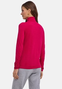 Dopasowany top z długim rękawem w jasnym różowym kolorze, z wysokim kołnierzem i miękką fakturą. Noszony z szarymi spodniami dresowymi.