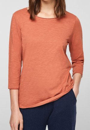 Kobieta ubrana w długi rękaw w kolorze rdzy z dekoltem łódkowym i ciemnoniebieskie spodnie, stojąca z jedną ręką w kieszeni na szarym tle.