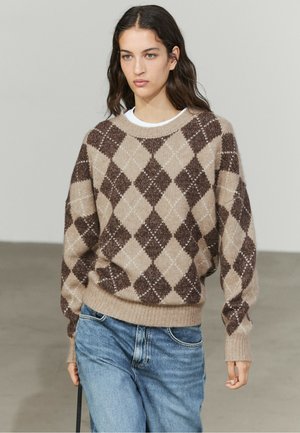 Ung kvinde iført en beige og brun argylemønstret sweater og blå jeans, der går indendørs mod en ensfarvet baggrund.