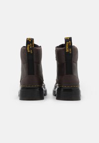 Botas de tobillo marrones con suelas de goma negras, con acentos amarillos, paneles laterales elásticos y un asa en la parte trasera para facilitar su uso.