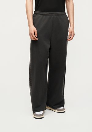 Osoba nosząca luźne czarne spodnie dresowe, czarny top, białe skarpetki i szare tenisówki, stojąca na tle jednolitego jasnego tła.