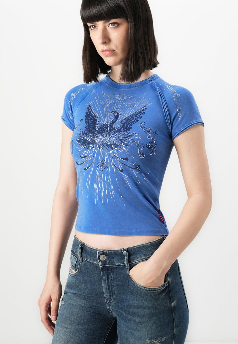 Woman with straight dark hair wears a fitted blue T-shirt with a black phoenix and studded design, paired with blue jeans, hand in pocket.