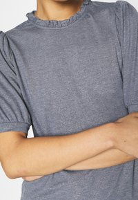 Top gris jaspeado con mangas cortas abullonadas y un escote con volantes. La tela tiene un brillo sutil, añadiendo textura.