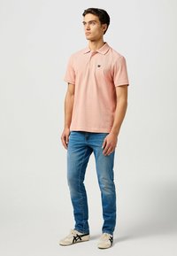 Polo shirt rosa chiaro con colletto, maniche corte e un piccolo logo. Abbinato a jeans blu e scarpe da ginnastica bianche con dettagli neri.