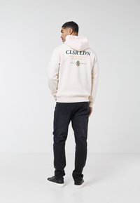 Man som bär en krämfärgad hoodie med texten CLSR LDN och emblem, svarta jeans och svarta sneakers, stående mot en enfärgad vit bakgrund.