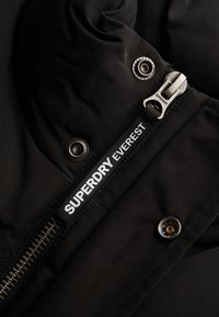 Close-up van een zwarte jasrits met "SUPERDRY EVEREST" branding en metalen drukkers op de stof nabij de ritssluiting.