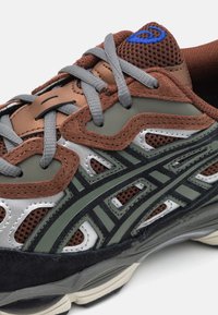 Baskets avec une tige en suède et en maille marron, des accents verts et argentés, des lacets gris, et une semelle en caoutchouc noir avec un motif.