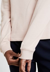 Felpa rosa chiaro con una texture soffice, polsini a coste e un logo bianco sulla manica. Abbinata a pantaloni blu scuro.