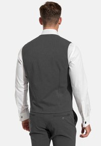 Gilet en tissu gris avec une coupe ajustée, présentant une texture lisse, deux fentes dans le dos et une fermeture à un bouton. Manches de chemise blanche visibles.
