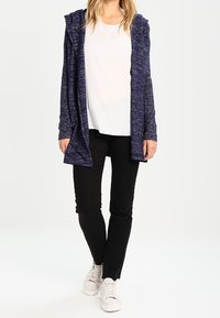Cardigan lungo in tessuto navy con cappuccio, design aperto davanti. Abbinato a una camicia bianca e pantaloni neri, completato con sneakers bianche.