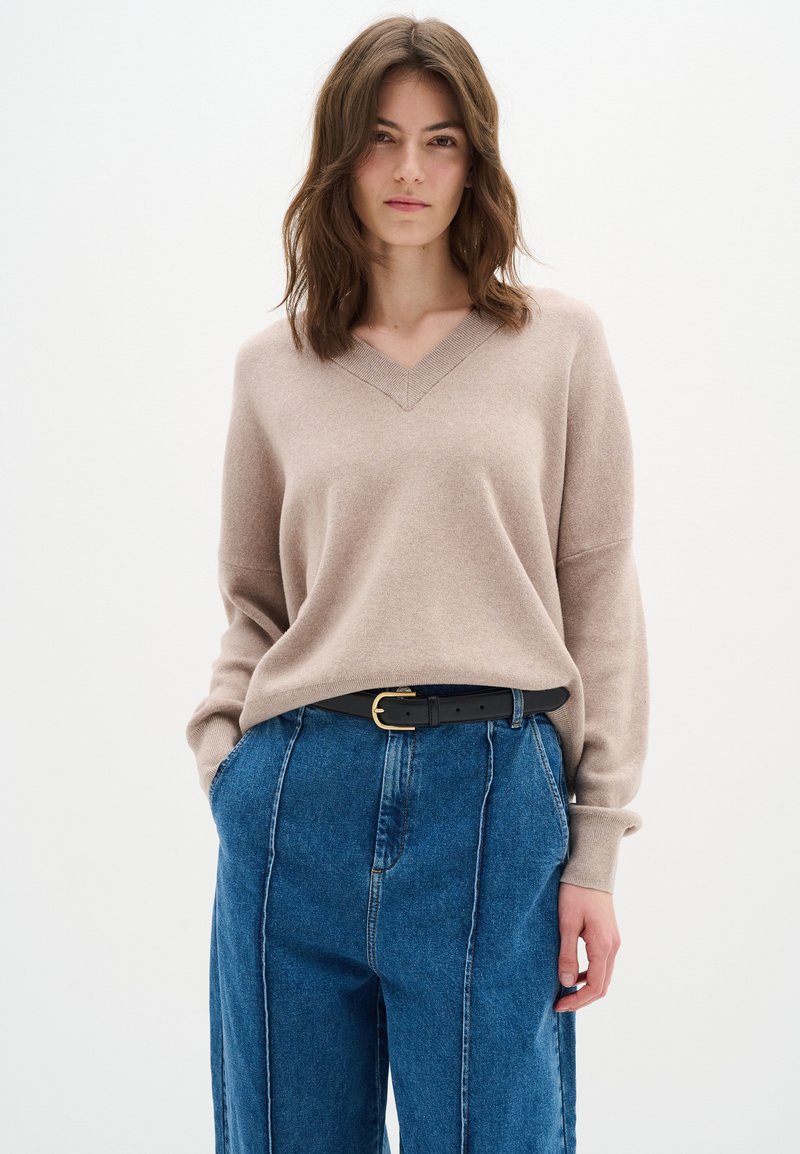 Beżowy sweter v-neck z długimi rękawami, w połączeniu z niebieskimi spodniami dżinsowymi z wysokim stanem, ozdobionymi pionowym szwem i czarnym paskiem.