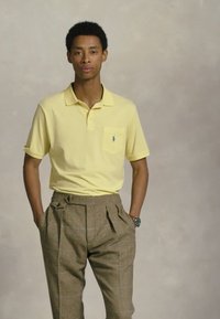 Moški v rumeni kratkosleeven polo majici in visokih rjavih kariranih hlačah, z rokama v žepih, stoji ob nevtralnem ozadju.