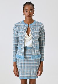 Cardigan a maglia a quadri blu e bianchi con dettagli frangiatati, abbinato a una gonna aderente. Il top è in satin crema, con scollatura a scialle.