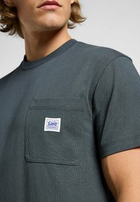 T-shirt verde scuro in cotone, con scollatura rotonda, maniche corte e taschino sul petto con etichetta bianca. Texture liscia.