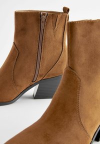 Stivali alla caviglia in suede marrone con zip laterale, tacco basso angolato e dettagli di cucitura sottili. Texture liscia e design snello.