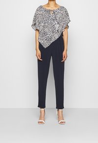 Blouse à motifs multicolores avec un col rond et un détail en forme de trou de serrure, associée à un pantalon bleu marine et des sandales à talons blancs.