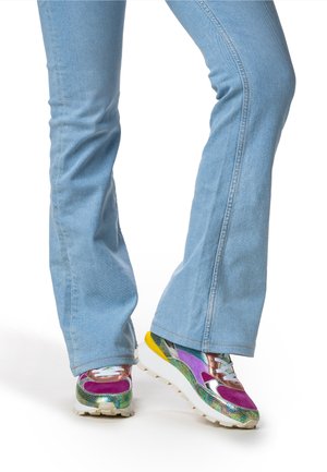 Sneakers piattaforma multicolore con un mix di texture, inclusi dettagli metallici, toni di viola, verde e giallo, abbinati a jeans a zampa d'elefante azzurri chiari.