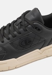 Schwarzer Ledersneaker mit gesticktem Champion „C“-Logo an der Seite, schwarzen Schnürsenkeln, gepolstertem Kragen und beiger Gummisohle.