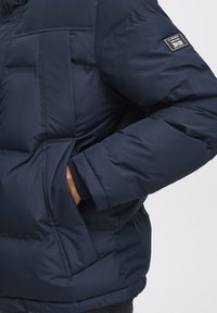 Jachetă puffer bleu marin cu un design matlasat, având o buzunare lateral, manșete ribbate și un eticheta din țesătură pe partea superioară a brațului.