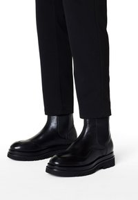 Botas de piel negras hasta el tobillo con un acabado brillante, puntera redonda, paneles laterales elásticos y suela de goma gruesa y texturizada. Combinadas con pantalones ajustados.