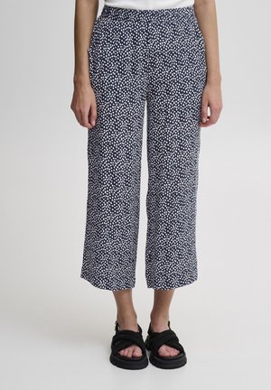 Frau trägt marineblaue kurze Hosen mit weißem verstreutem Punktmuster und schwarzen klobigen Sandalen mit Riemen.