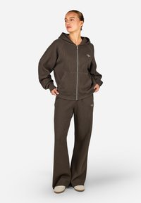 Hnědá žebrovaná mikina s kapucí na předním zipu, velkými kapsami a výšivkou, doplněná o sladěné žebrované kalhoty s volným střihem. Měkká textura.