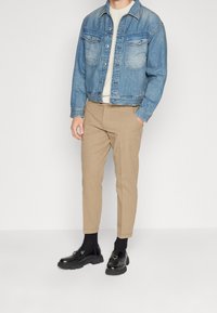Giacca in denim blu, maglione lavorato a maglia beige, pantaloni beige, mocassini neri, con materiali testurizzati e una vestibilità su misura. Due tasche sul petto della giacca.