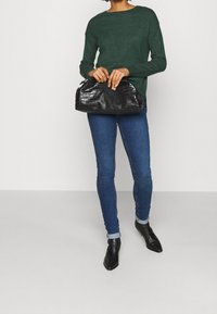 Grön långärmad tröja, mörkblå smaljeans, svarta spetsiga ankelskor, hållande en svart handväska i krokodilskinn. Neutral bakgrund.