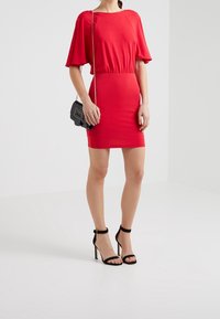 Kobieta w czerwonej, krótkiej sukience z falbanami, czarnych szpilkach oraz z małą czarną torebką na ramię z złotym łańcuszkiem.