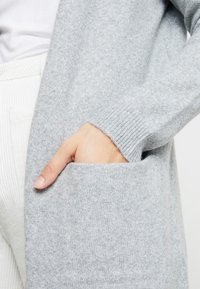 Hellgrauer Cardigan aus weichem Strickmaterial mit einer Tasche auf der rechten Seite. Mit gerippten Bündchen und einem offenen Frontdesign.